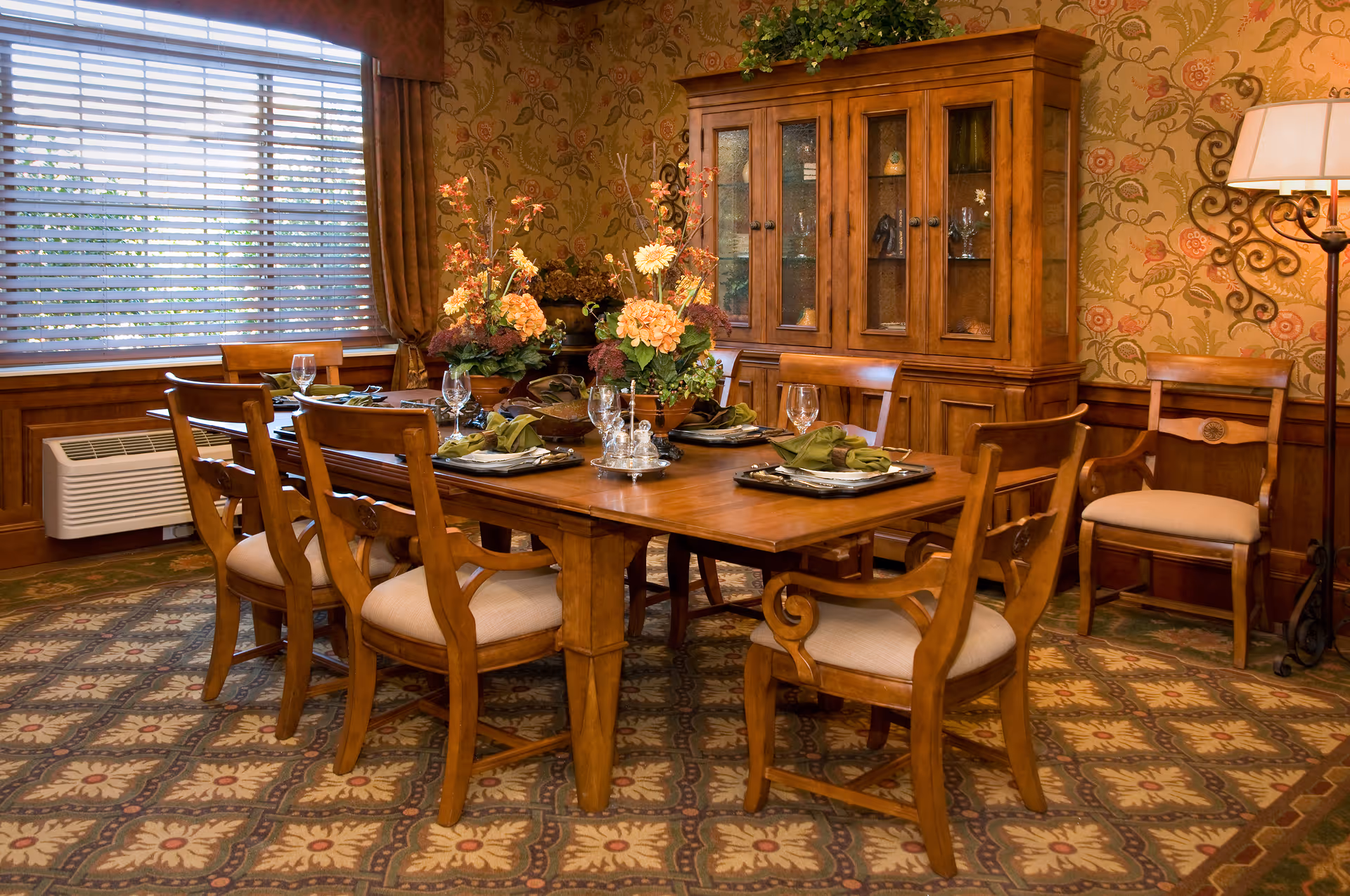 A warmly decorated dining room with a wooden dining table set for six, featuring floral centerpieces, glassware, and folded napkins. The room has patterned wallpaper, a large window with blinds, a wooden china cabinet, and a floor lamp in the corner.