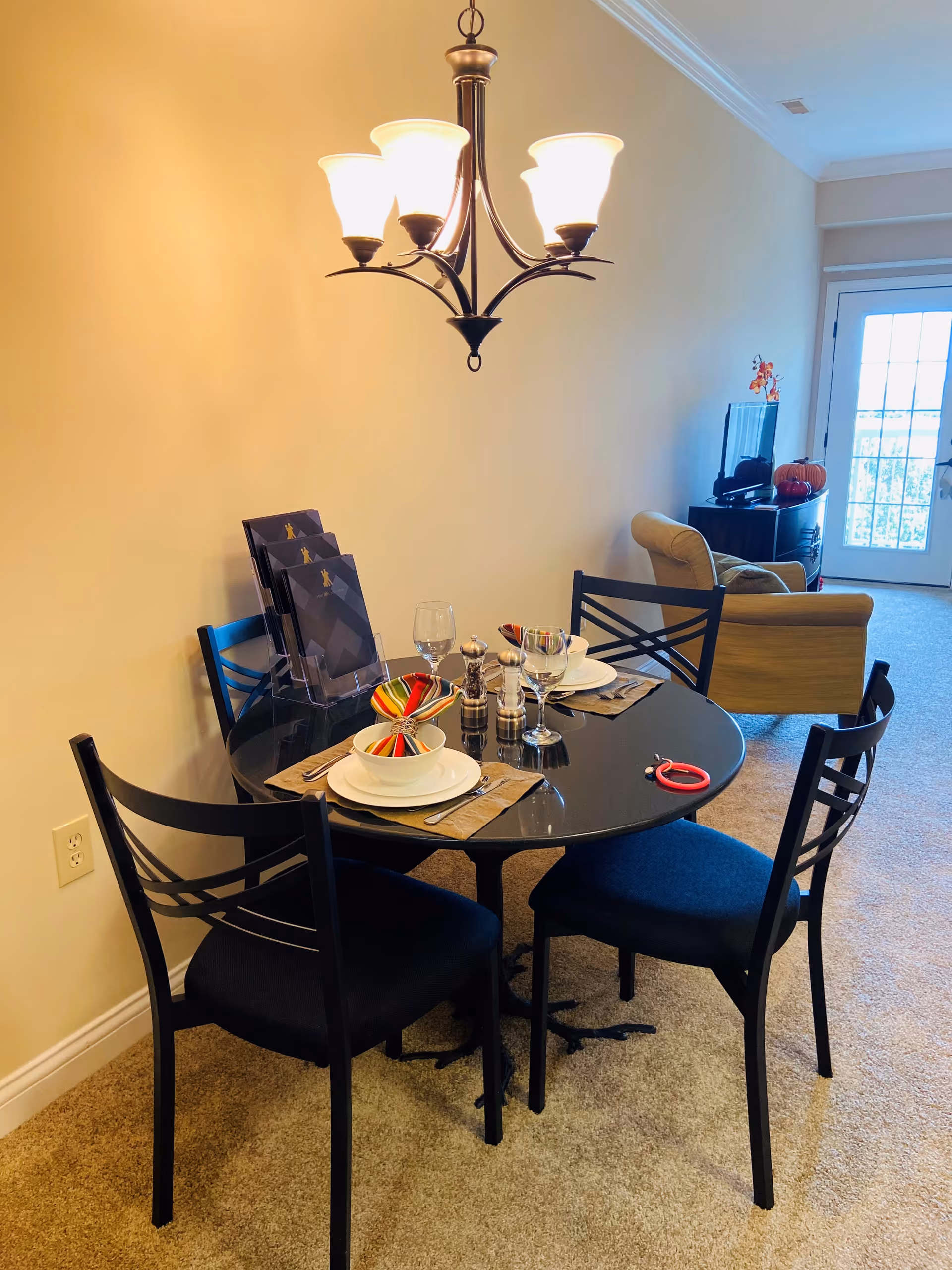 Round black dining table with place settings and four chairs under a chandelier in a living area near a glass door.
