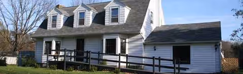 White two-story house with a wooden ramp, dormer windows, and surrounding lawn.