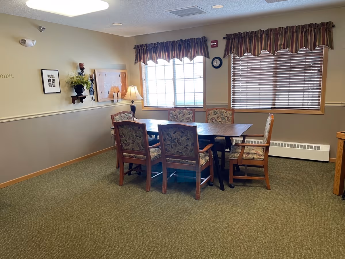 A small meeting or dining room with a rectangular wooden table surrounded by six cushioned chairs with floral upholstery. The room has beige walls with a chair rail, green carpet, two windows with blinds and valances, a wall clock, a lamp on a small table, and some wall decorations including a framed picture and a bulletin board.