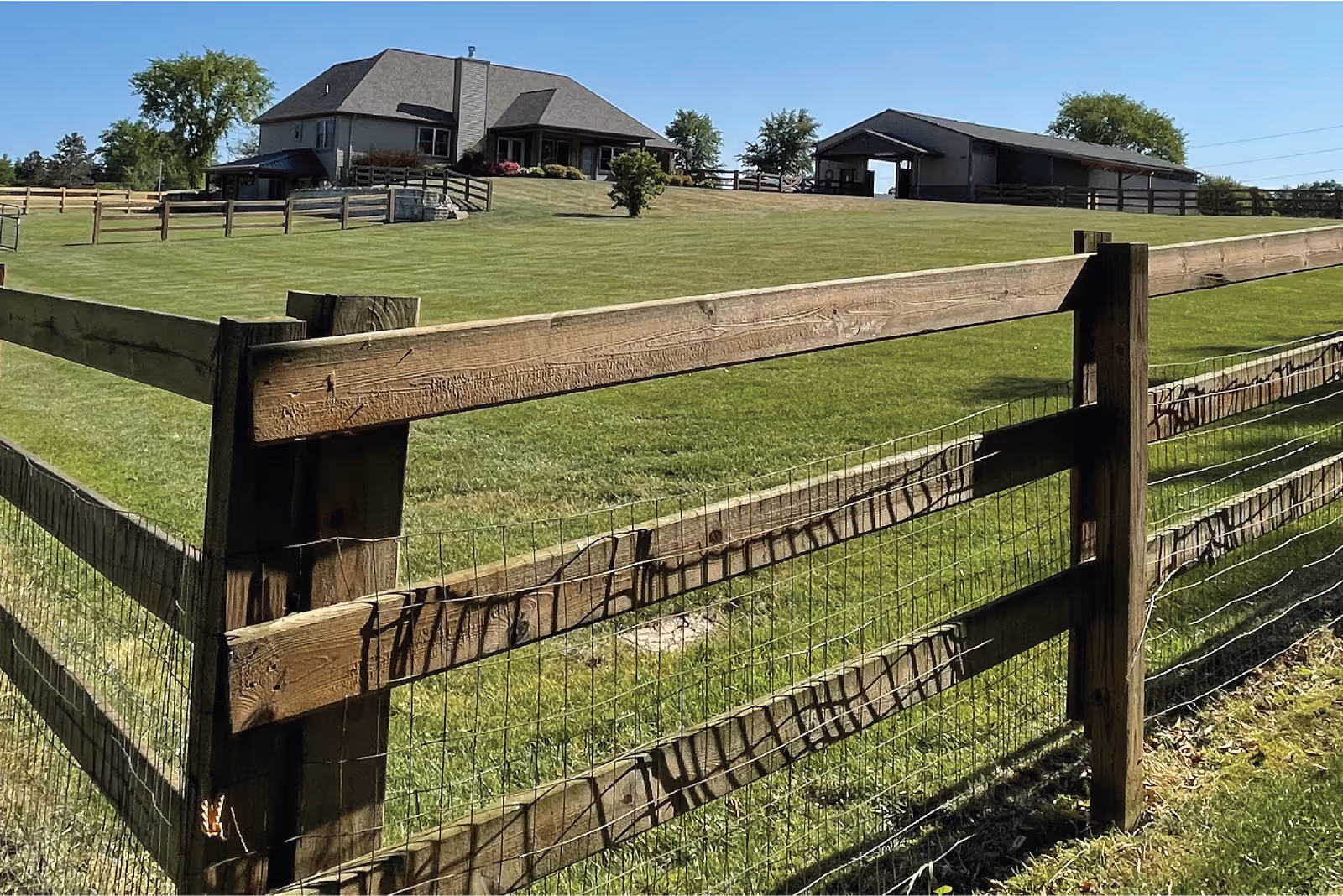 Wooden fence and green lawn leading up to a house and a barn under a clear blue sky.