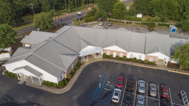 Aerial view of a single-story building with a gray roof and white walls, surrounded by greenery and a parking lot with several parked cars. The building has a covered entrance and is located near a road with trees and bushes around the property.