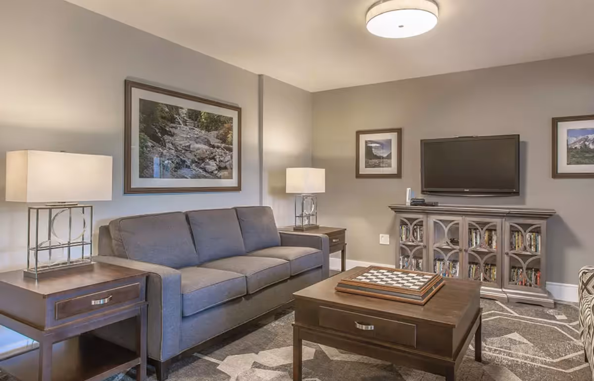 Living room with a gray sofa, wooden coffee table with a chessboard, TV on a cabinet, lamps, and framed artwork.