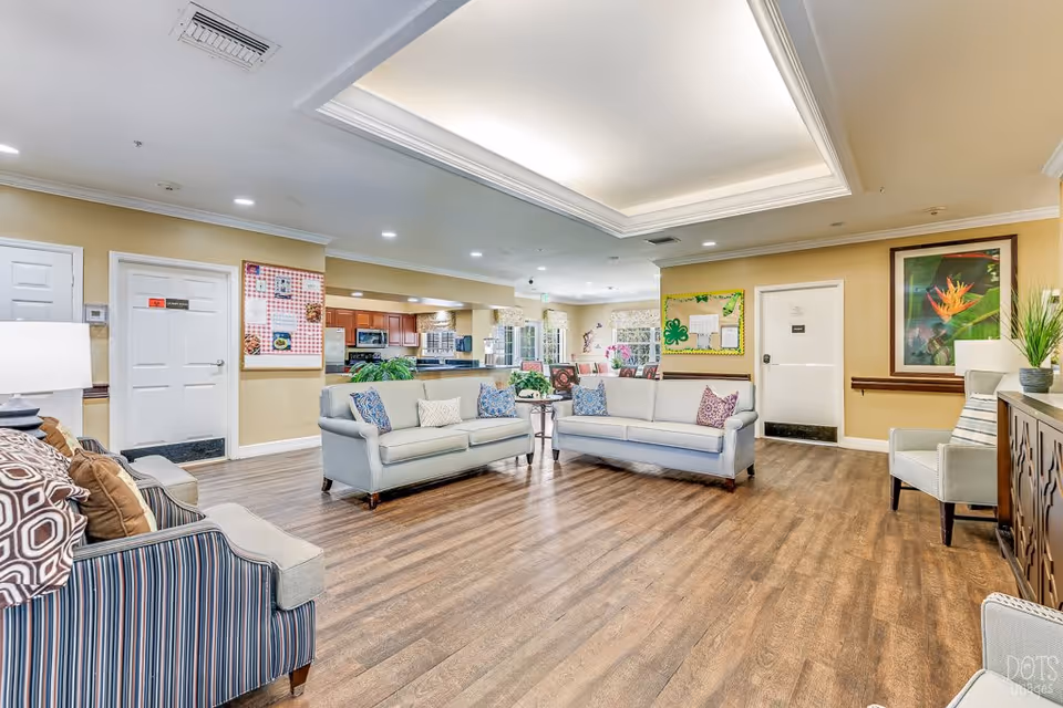 A spacious and well-lit living room area in a senior living facility featuring multiple sofas and armchairs with decorative pillows arranged around a central coffee table. The room has wood flooring, beige walls, recessed ceiling lighting, and framed artwork on the walls. There is a kitchen area visible in the background with wooden cabinets and stainless steel appliances.