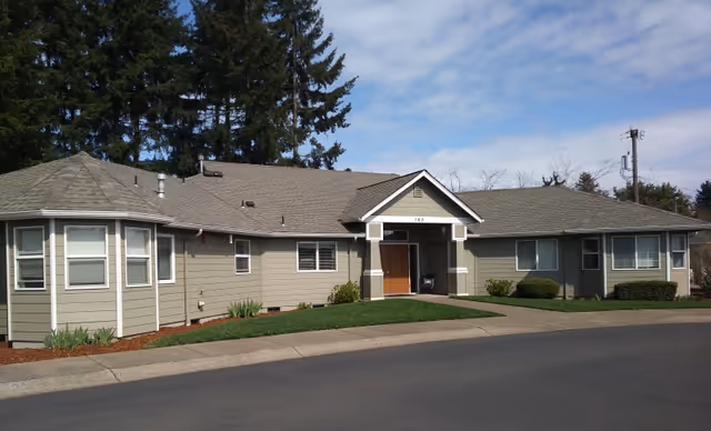 Single-story River Grove Memory Care exterior with beige siding, front entrance, and surrounding greenery.