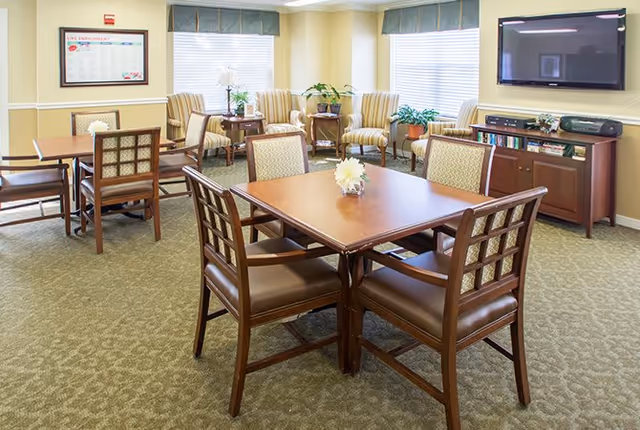 A bright and cozy common area in a senior living facility featuring several wooden tables with cushioned chairs arranged around them. The room has large windows with blinds and valances, allowing natural light to fill the space. There are two striped armchairs and a small table with a lamp and plants near the windows. A flat-screen TV is mounted on the wall above a wooden cabinet that holds books and a stereo system. The carpet is patterned in a neutral tone, and a framed schedule or notice is hung on the wall.