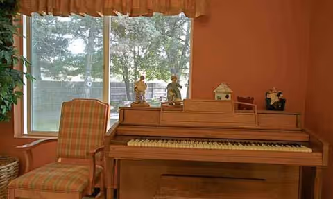 A wooden piano with figurines sits in a pink-walled living room beside a plaid chair