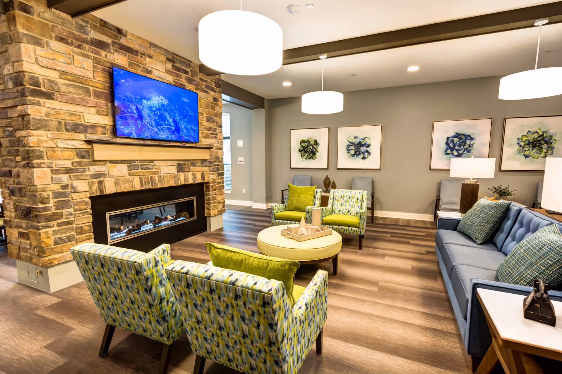 A cozy senior living facility lounge with a stone fireplace and a mounted flat-screen TV above it. The room features a blue sofa with green and blue patterned pillows, two patterned armchairs with green cushions, a round ottoman coffee table, and additional seating along the back wall. The walls are decorated with framed botanical artwork, and modern pendant lights hang from the ceiling, illuminating the space.