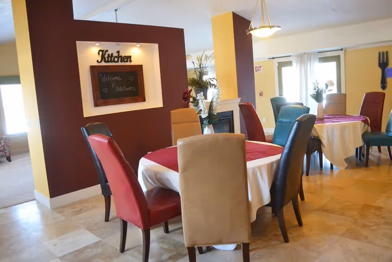 Bright dining room with round tables covered in white and red cloths, upholstered chairs, and a wall niche sign reading "Kitchen".