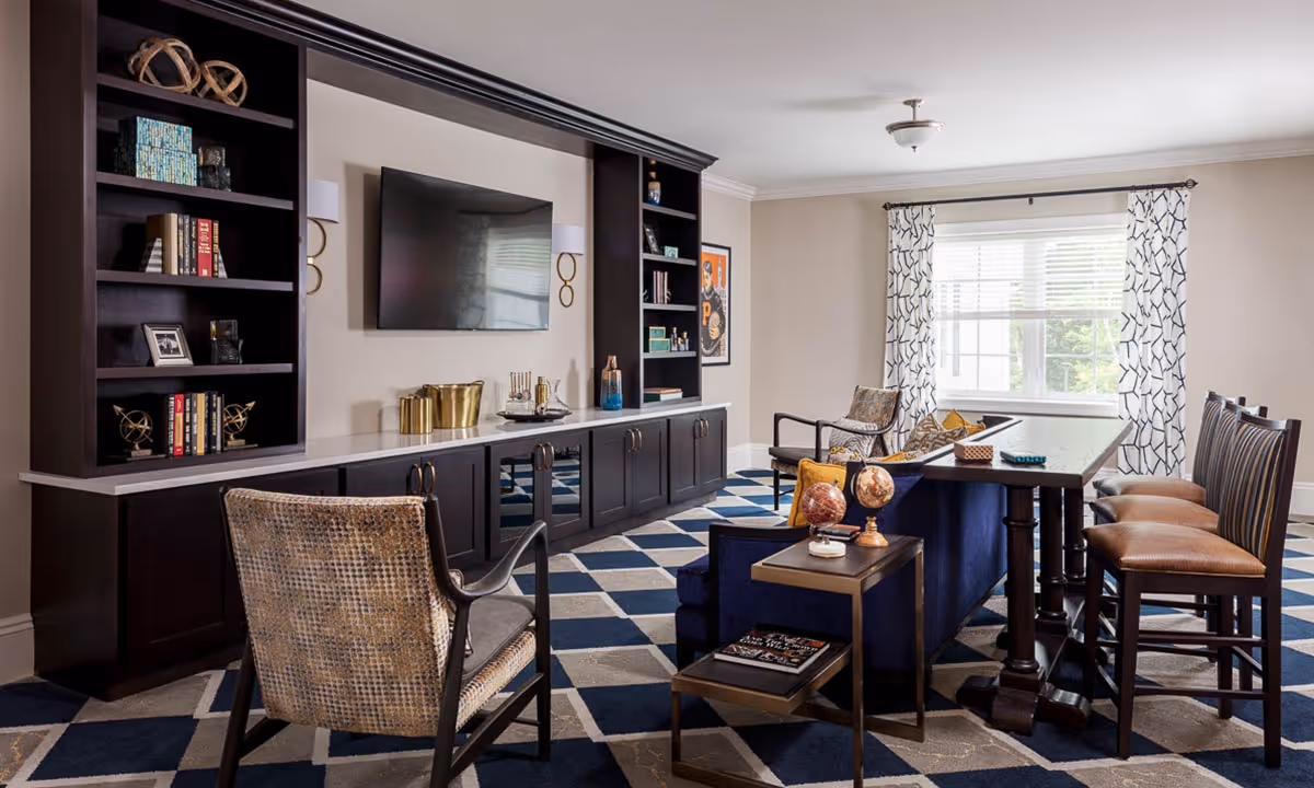 Communal living room/lounge with dark built-in shelving and mounted TV, seating, a high table with stools, and a large window with patterned curtains.