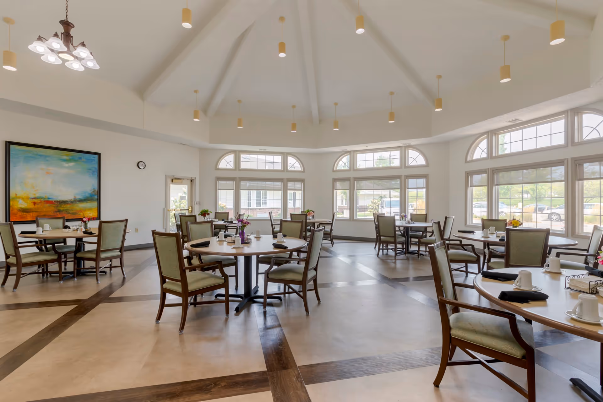 Bright senior living dining room with round tables and chairs, large windows letting in natural light, a high ceiling with pendant lights, and a colorful abstract painting on the wall.