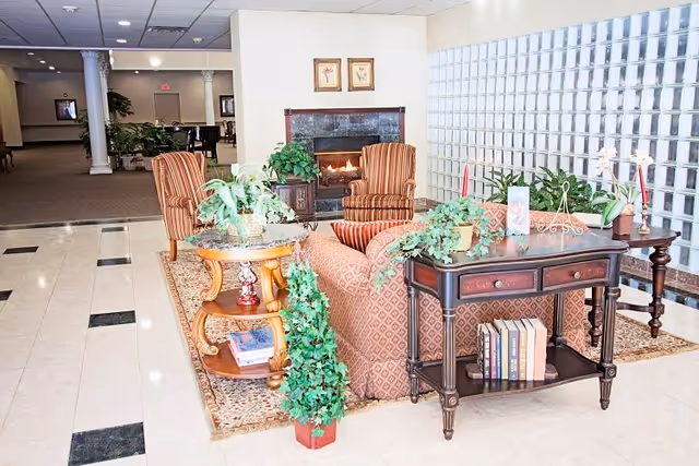 A cozy seating area in a senior living facility featuring a patterned sofa and two striped armchairs arranged around a fireplace. The space includes a wooden side table with books and decorative plants, a dark wood console table with more books and plants, and a glass block wall providing natural light. The floor is tiled with a beige and black pattern, and the area is well-lit with ceiling lights.