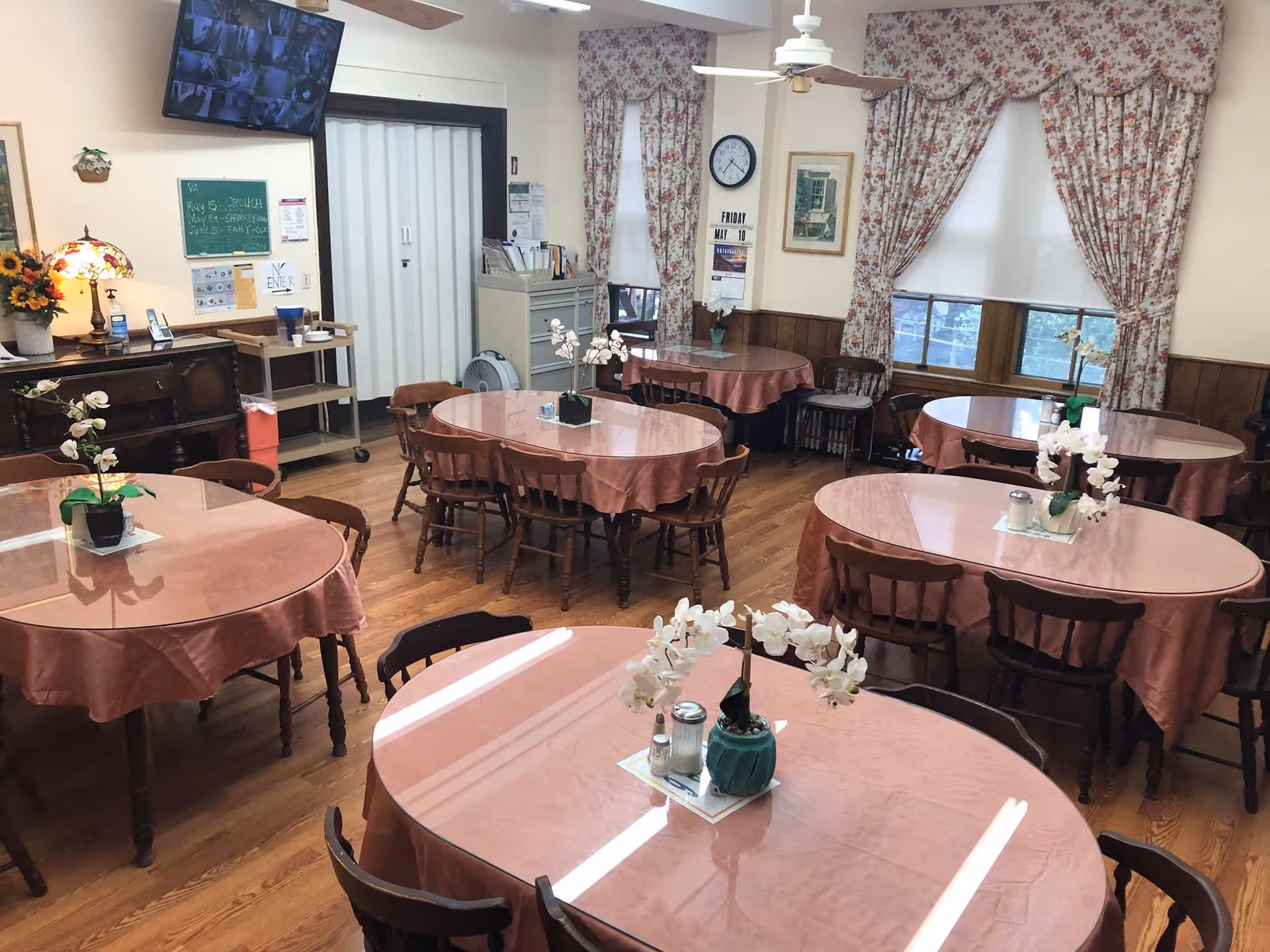 Communal dining room with multiple round tables draped in pink tablecloths, wooden chairs, floral centerpieces, and floral curtains.