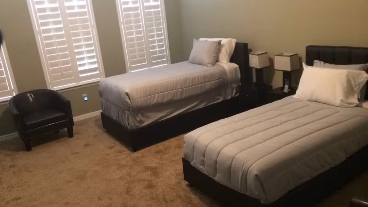 A bedroom with two single beds, each with gray bedding and white pillows. Between the beds is a nightstand with two matching lamps. There is a black armchair in the corner near three windows with white shutters. The room has beige carpet and light-colored walls.