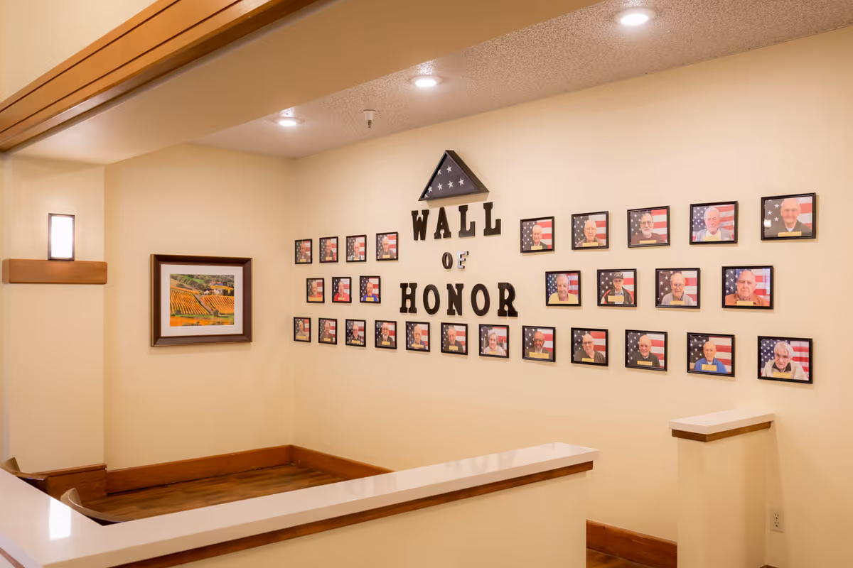 Interior view of a hallway corner in Cogir of Vacaville featuring a Wall of Honor with framed photos of individuals in front of American flags. The wall has large black letters spelling out 'WALL OF HONOR' and a folded American flag displayed above the text. There is also a framed picture of a vineyard on the adjacent wall.