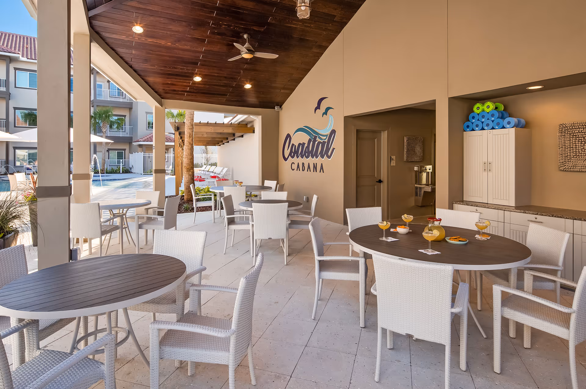 Outdoor covered seating area with round tables and white chairs, a wooden ceiling with ceiling fans, and a wall with a 'Coastal Cabana' sign. In the background, there is a swimming pool area with lounge chairs and umbrellas.