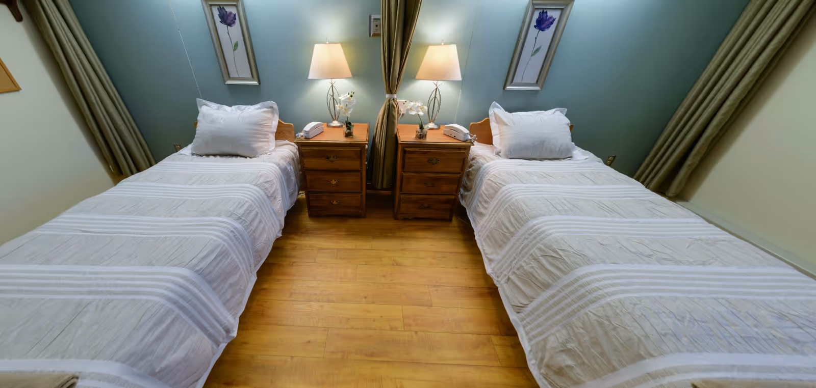 A bedroom with two single beds separated by two wooden nightstands, each with a lamp, a telephone, and a small flower arrangement. The beds have white bedding and pillows. The walls are painted light blue and decorated with framed floral artwork. There are green curtains on either side of the beds.