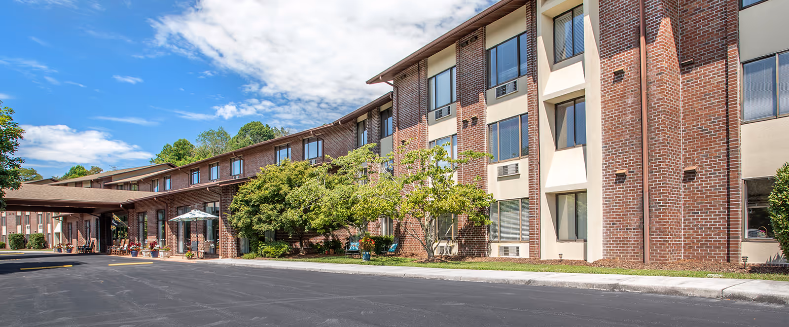 Exterior view of a multi-story brick building with large windows, a covered entrance, and landscaping including trees and potted plants under a partly cloudy blue sky.