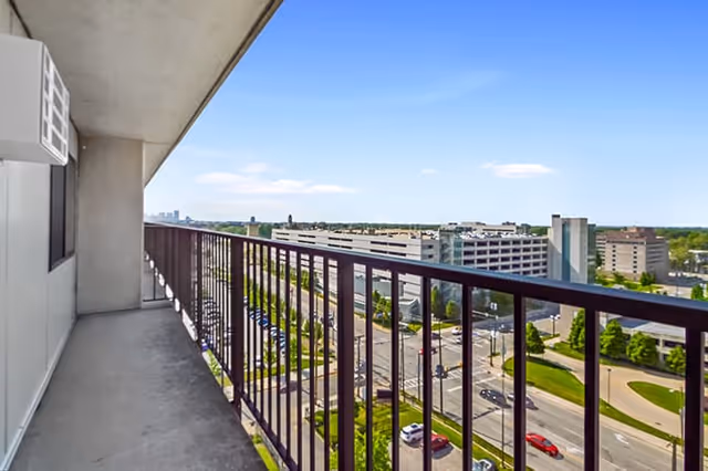 High-rise balcony with a metal railing overlooking parking garages, streets and buildings under a clear blue sky.
