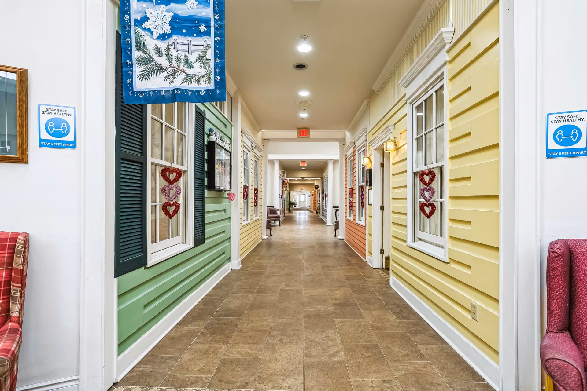 A brightly lit hallway in a senior living facility with walls designed to look like colorful house exteriors in green, yellow, and orange. The hallway has tiled flooring and is decorated with heart-shaped ornaments on the windows. There are signs on the walls reminding to stay safe and stay healthy by maintaining 6 feet apart. Chairs with red plaid and solid red upholstery are partially visible on either side of the hallway.