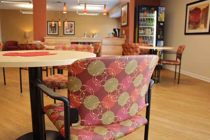 Interior view of a common dining area with round tables and patterned chairs. The room has wooden flooring, warm lighting with hanging pendant lights, framed artwork on the walls, and a vending machine in the background.