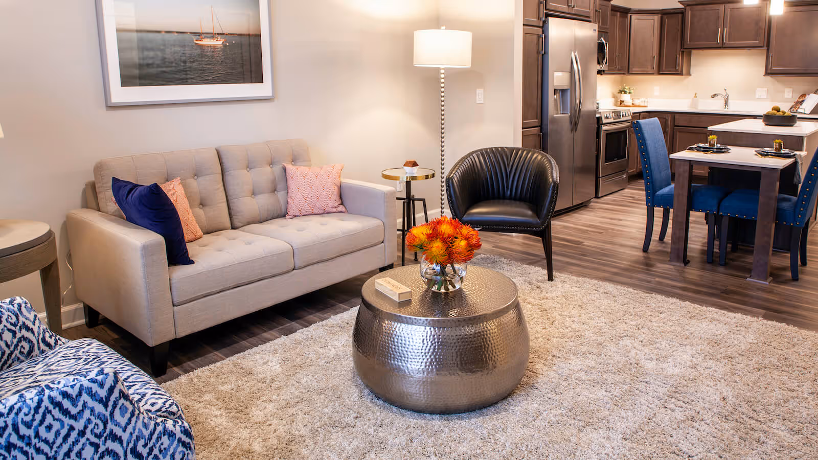 Modern open-plan living room with a beige sofa, black accent chair, metallic coffee table with flowers, and a view into a kitchen and dining area.