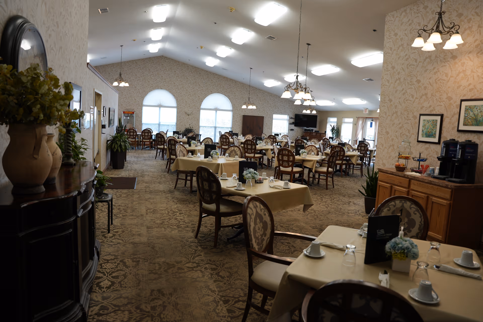 A spacious dining room with multiple tables covered in beige tablecloths, each set with cups, glasses, and napkins. The room has patterned carpet, floral wallpaper, and several chandeliers hanging from the ceiling. There are large arched windows at the far end letting in natural light, and a sideboard with coffee machines and a juice dispenser on the right side.