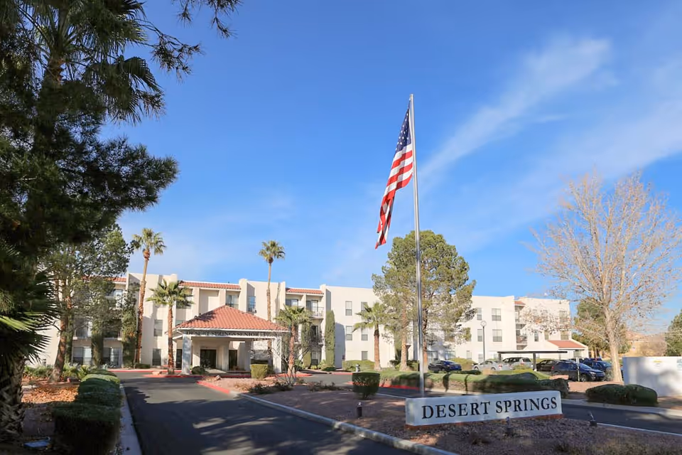 Front entrance of the Desert Springs senior living building with an American flag on a tall flagpole and a sign reading "DESERT SPRINGS".