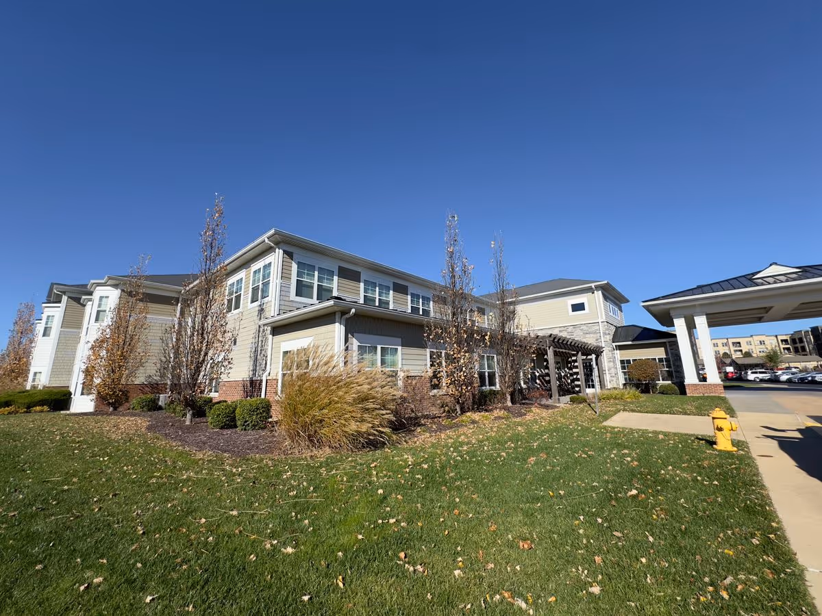 Exterior view of a senior living building with a covered entrance, landscaped lawn, and clear blue sky.