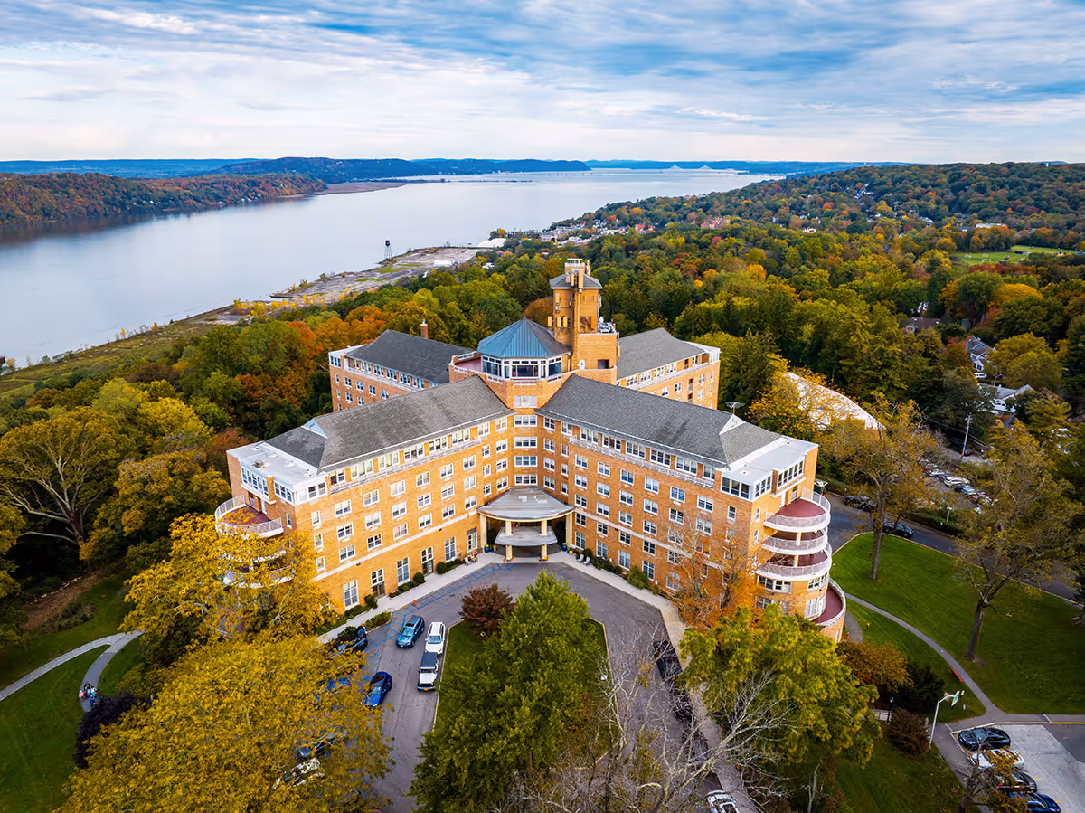 Aerial view of a large multi-story brick senior living building with a circular driveway, surrounded by trees and a river in the background.
