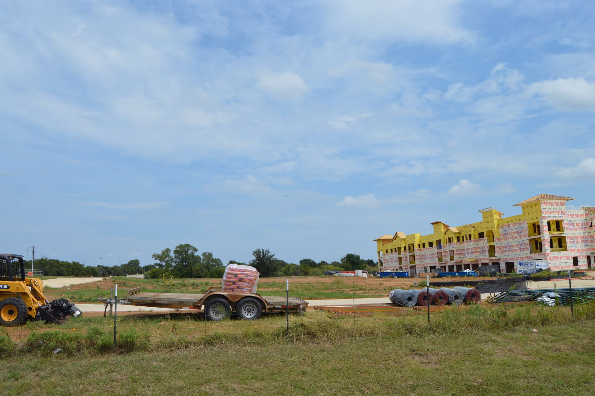 Construction site with a partially built multi-story building wrapped in yellow insulation material. In the foreground, there is a trailer loaded with construction materials and a small construction vehicle. The area is grassy with some fencing, and the sky is partly cloudy.