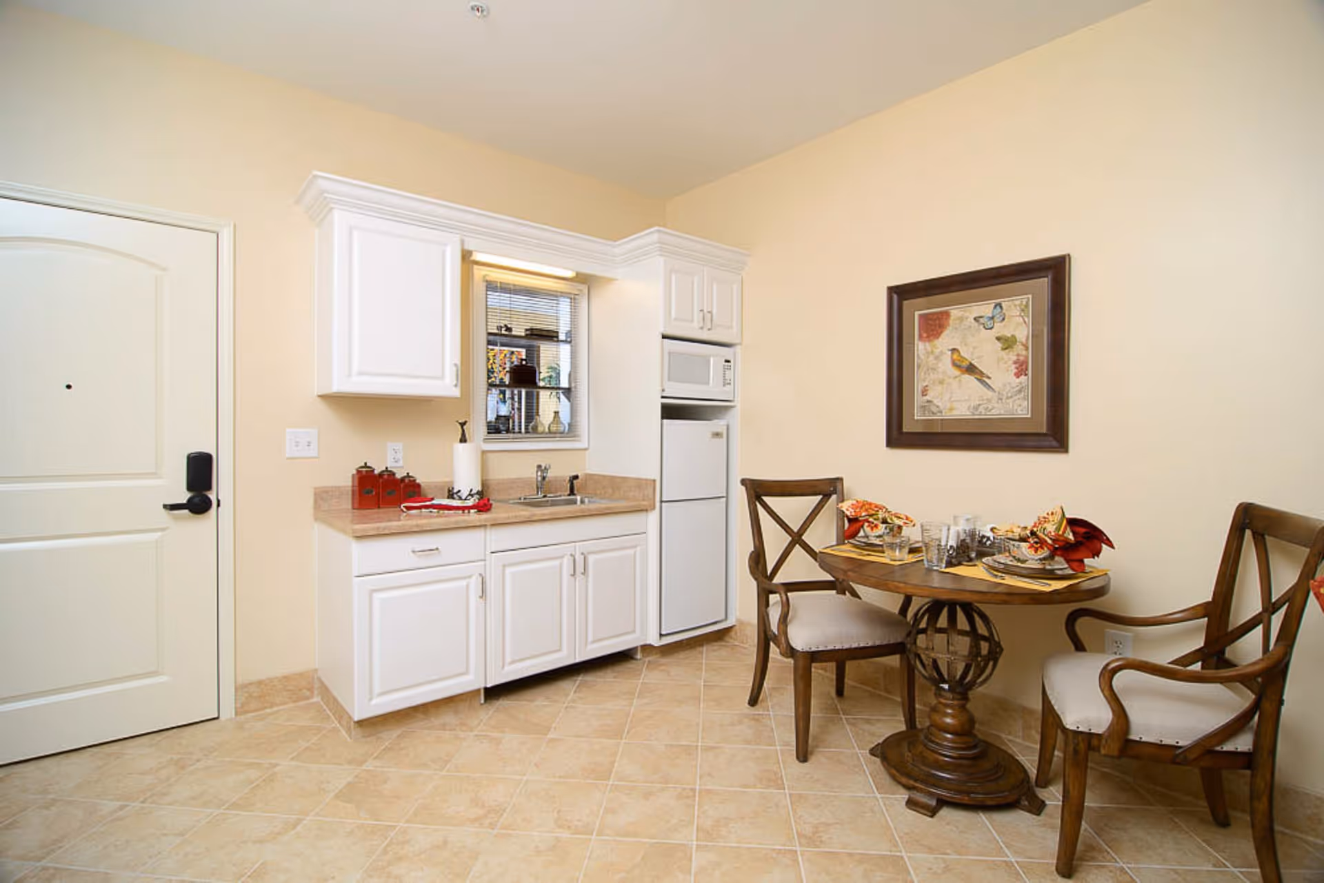 Small kitchen area with white cabinets, a countertop with a sink, a microwave, and a mini refrigerator. Next to the kitchen is a round wooden dining table set for two with two wooden chairs. A framed picture of a bird and butterfly hangs on the wall above the table. The floor is tiled and the walls are painted light beige.