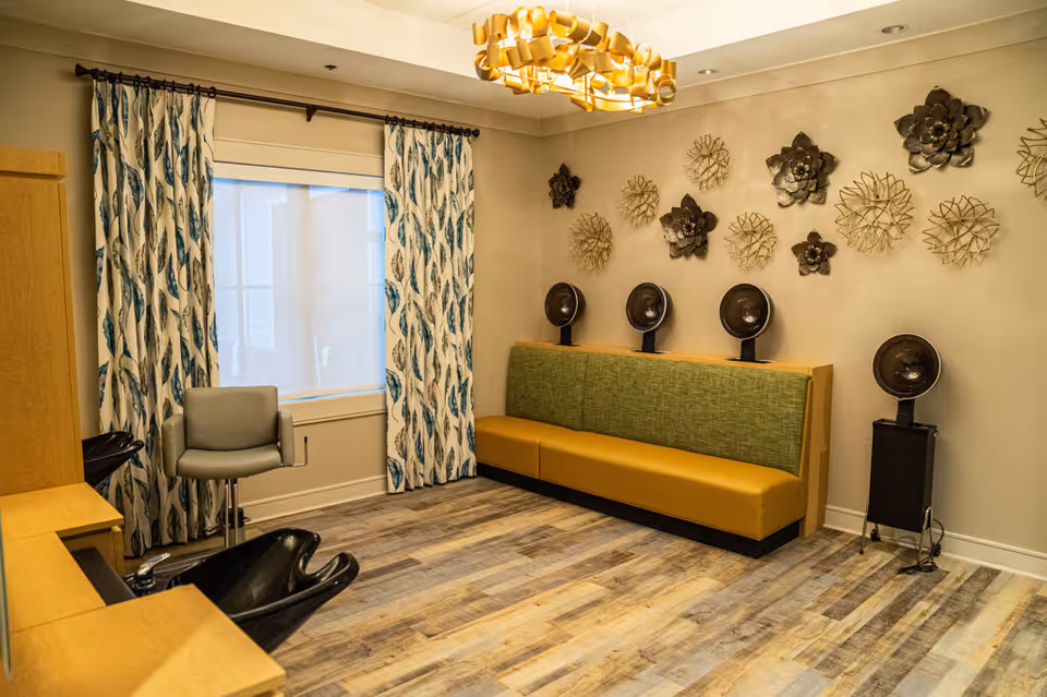 Interior of a salon room with a green and yellow cushioned bench along one wall, five black hair dryer chairs mounted on the wall and one standing, a gray salon chair, wooden cabinetry, patterned curtains with blue and white leaf designs, and decorative metal wall art shaped like flowers.