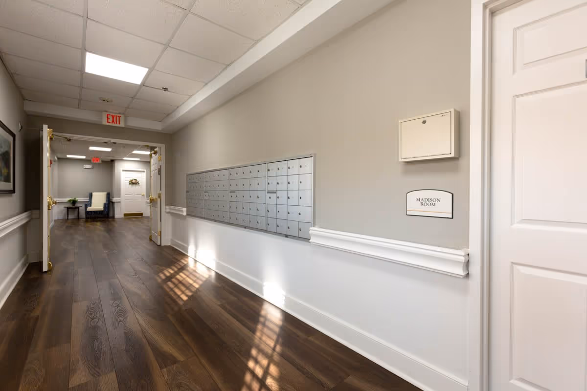 Interior hallway of a senior living facility with wooden flooring, a row of mailboxes mounted on the right wall, and an open doorway leading to a room with a chair and table. The walls are painted light gray with white trim, and there is a sign on the wall labeled 'Madison Room'.