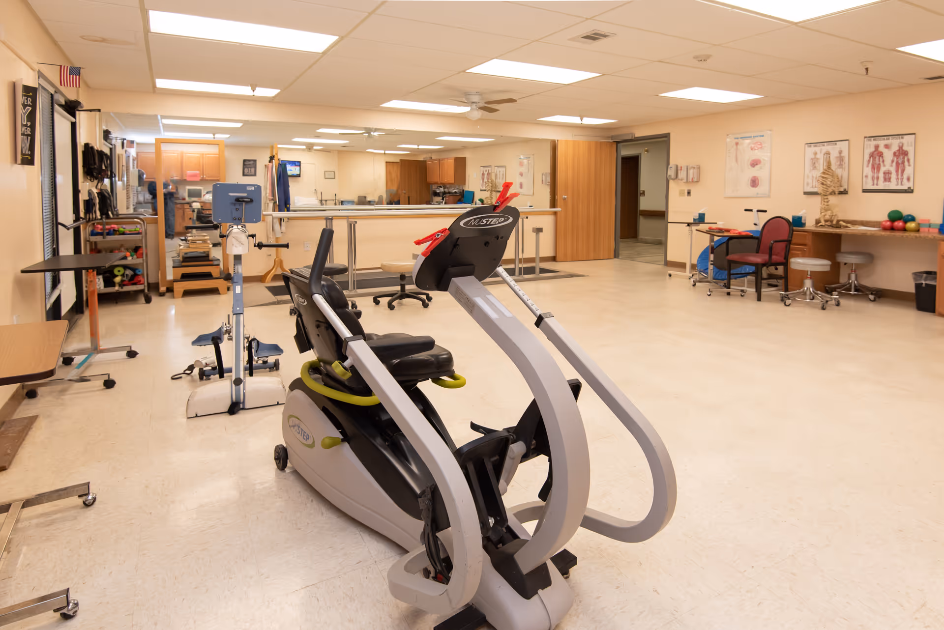A spacious physical therapy or rehabilitation room with exercise equipment including a NuStep recumbent cross trainer and a stationary bike. The room has beige walls, a large mirror on one side, anatomical posters on the wall, a skeleton model, and various therapy tools and chairs. The ceiling has fluorescent lighting and a ceiling fan.