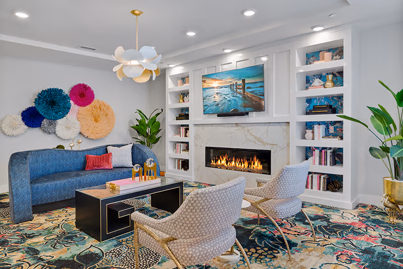 A modern living room with a blue curved sofa, two patterned armchairs, a black coffee table, and a colorful patterned rug. The room features a built-in white shelving unit with books and decorative items, a wall-mounted TV above a marble fireplace with a fire burning, and a unique ceiling light fixture. There are also large green plants and colorful circular wall decorations.