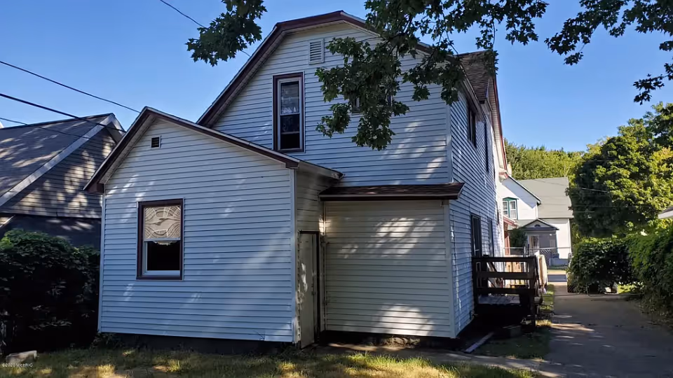 White two-story house with vinyl siding, a small side porch and driveway shaded by trees.