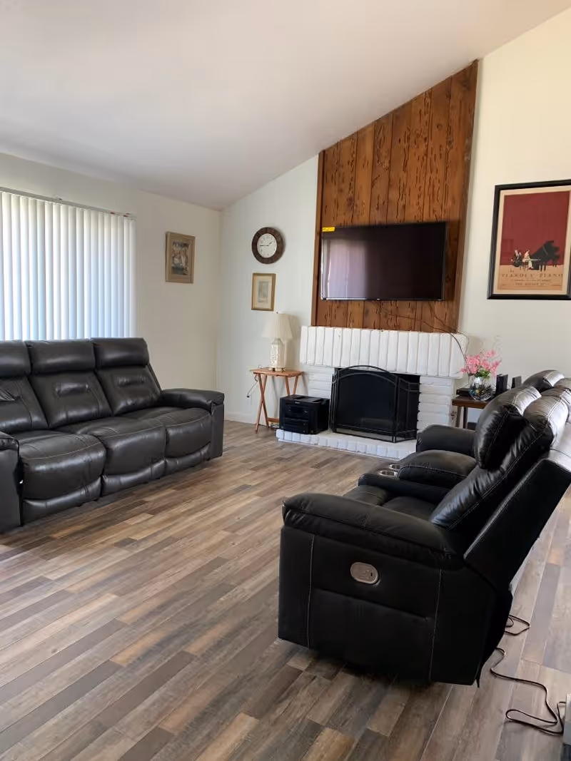 A living room with two black leather recliner sofas facing a white brick fireplace with a mounted flat-screen TV above it. The fireplace has a wooden panel backdrop. There is a small side table with a lamp and a clock on the wall, along with framed pictures. The floor is wood laminate, and vertical blinds cover a window on the left side.