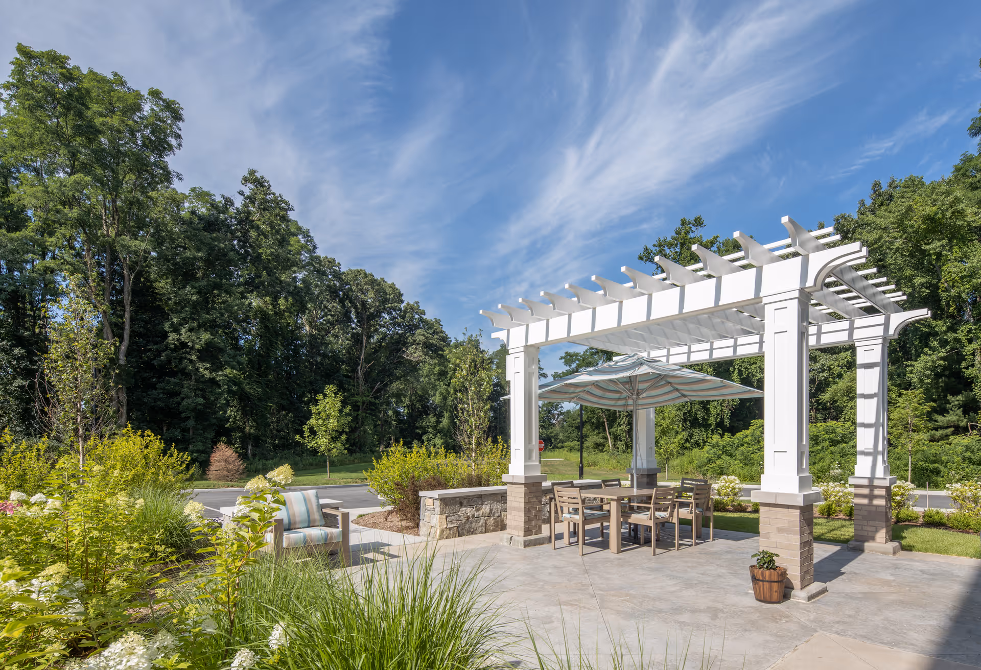 Outdoor patio area with a white pergola covering a table and chairs. There is a large umbrella over the table and a cushioned chair nearby. The patio is surrounded by greenery and trees under a partly cloudy blue sky.
