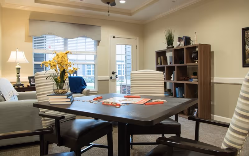 A cozy common area with a dark wooden table and four striped cushioned chairs. On the table, there is a vase with yellow flowers, some books, and a Scrabble game in progress. In the background, there is a window with blinds, a door leading outside, a bookshelf with various items, and a lamp on a side table next to a sofa.