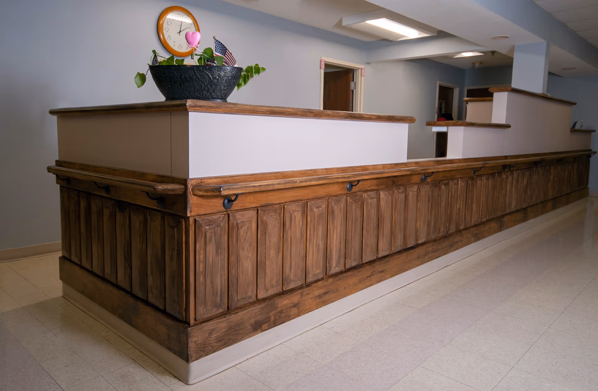 A long wooden reception or nurse station counter with a handrail along the front, topped with a plant in a black pot, a clock on the wall, and an American flag. The area has light-colored tiled flooring and light blue walls with doorways in the background.