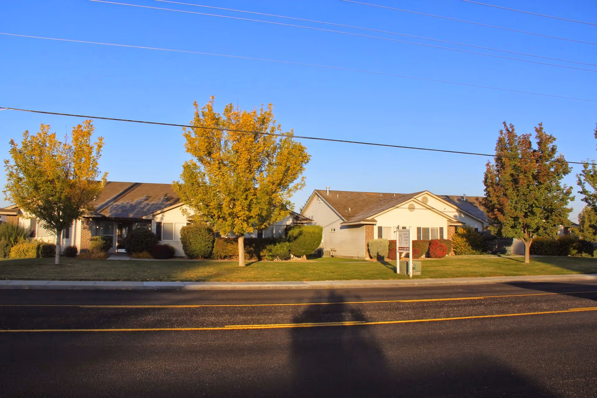 Exterior view of a single-story residential building with a brown roof and light-colored walls, surrounded by green grass, bushes, and several trees with autumn foliage under a clear blue sky.