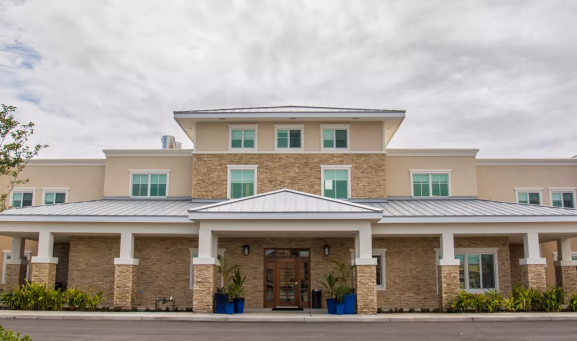 Front exterior view of a senior living facility building with beige and stone facade, multiple windows, a metal roof, and a covered entrance with blue potted plants on either side of the door.
