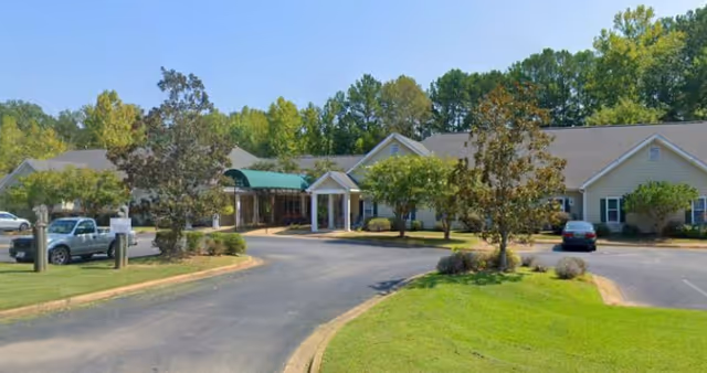 Single-story assisted living building with a covered entrance canopy, circular driveway, parked cars and landscaped lawn.