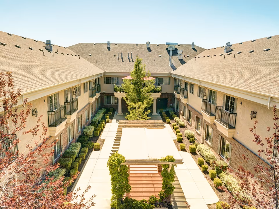 View of a courtyard in a senior living facility with a pergola covered in greenery in the center, surrounded by neatly trimmed bushes and trees, and two-story buildings with balconies on either side under a clear sky.