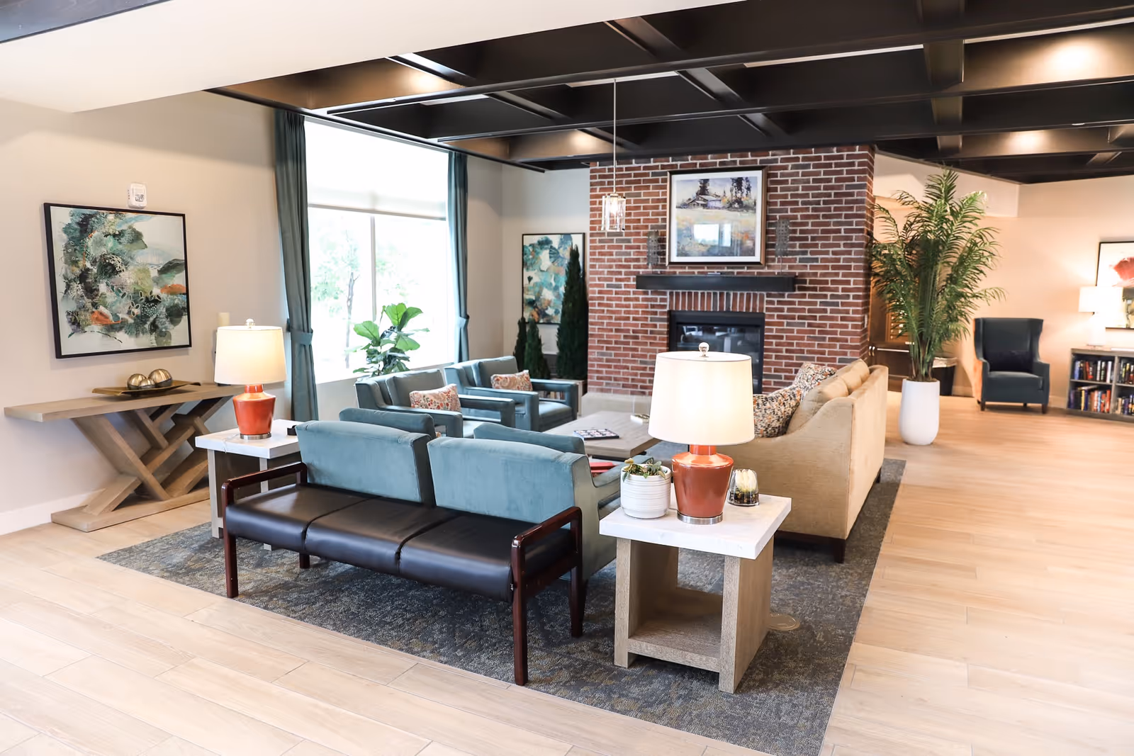 A spacious and well-lit living room area with a brick fireplace as the focal point. The room features a beige sofa, two teal armchairs, a black bench, and side tables with lamps and plants. There are large windows with teal curtains, abstract artwork on the walls, a large potted plant, and a bookshelf in the background.