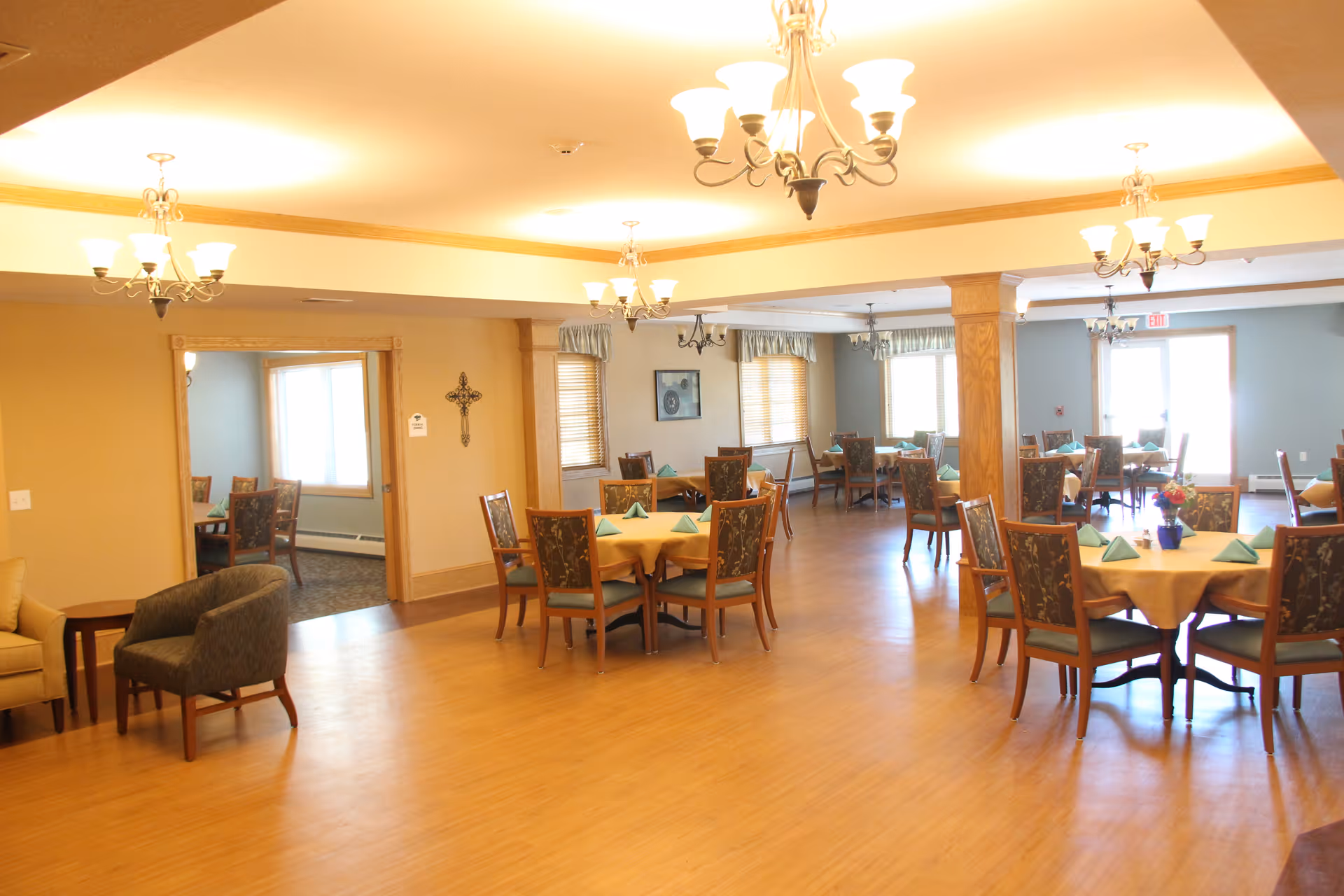 Spacious dining room with round tables set with napkins, upholstered chairs, chandeliers, and wood flooring.