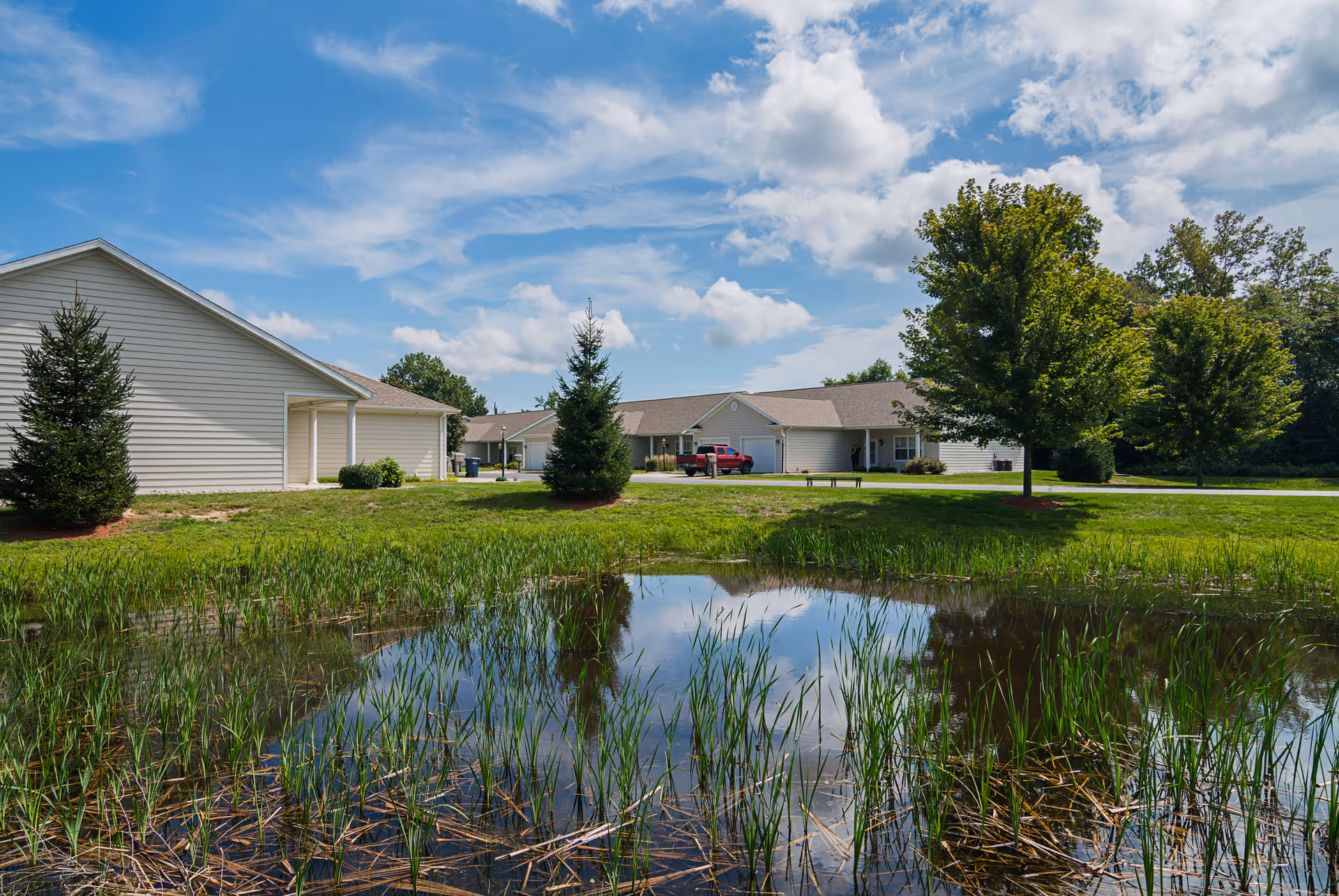 Exterior view of a senior living facility with single-story buildings, a small pond with tall grass in the foreground, green trees, and a partly cloudy blue sky.