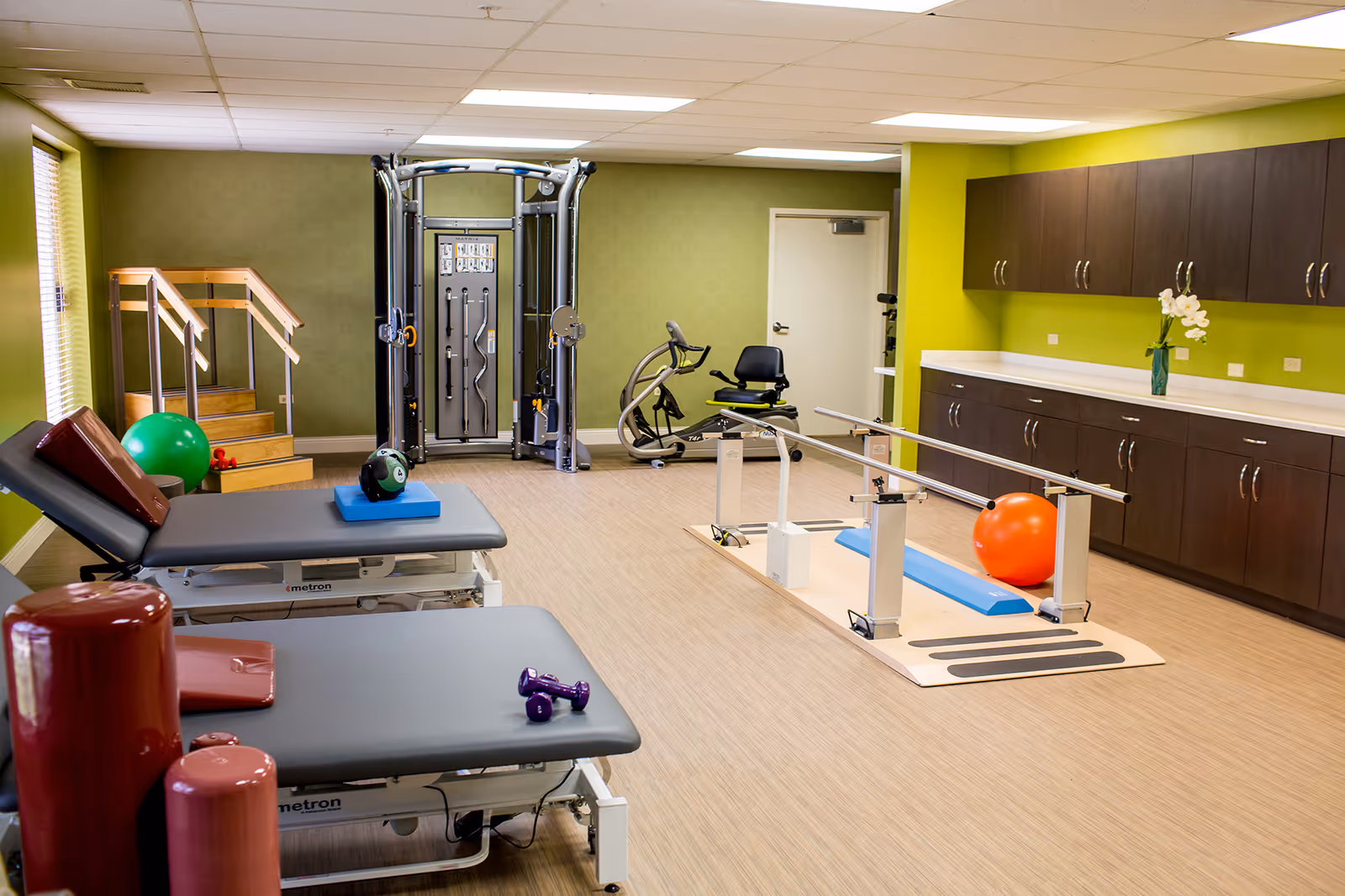 Physical therapy/exercise room with treatment tables, parallel bars, various fitness equipment, and cabinetry.