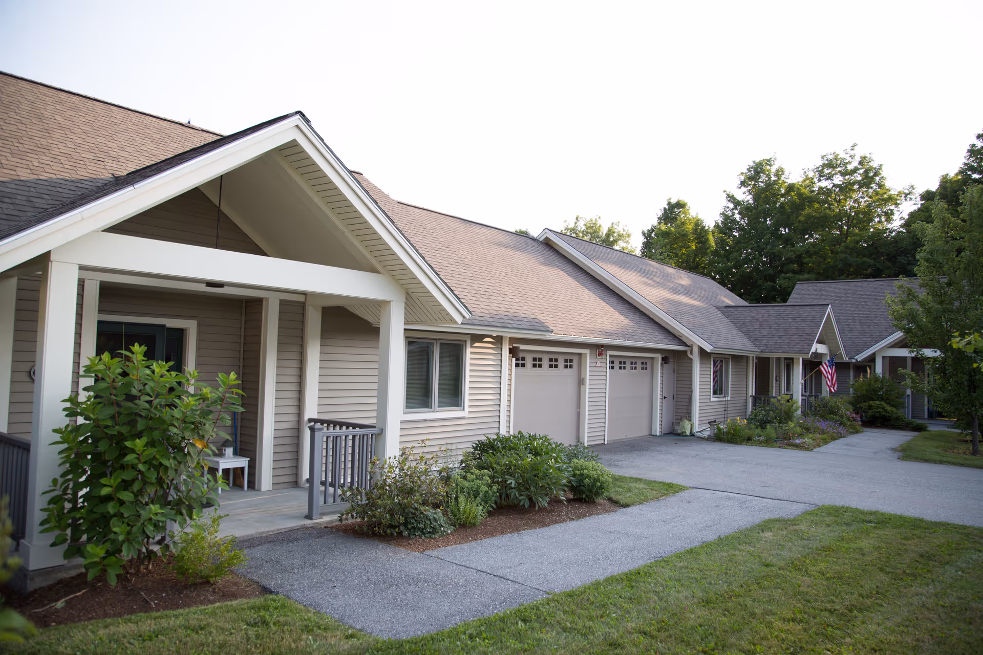 Exterior view of a single-story residential building with beige siding and a brown roof, featuring a small porch, garage doors, and landscaped greenery along the walkway. Trees and a well-maintained lawn surround the building.